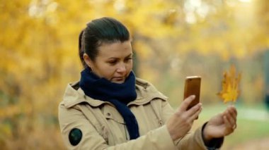 Genç bir kız telefonuyla düşen bir sonbahar akçaağaç yaprağının fotoğrafını çekiyor. İnsanlar, teknoloji ve doğa. Sonbahar günü. Yüksek kalite 4k görüntü