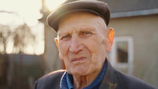 家の裏側の通りに立っている帽子を被った老人の肖像 カメラを近くで見ている老人の古いしわの顔 高品質の4K映像 — ストック動画