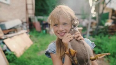 Küçük mavi gözlü sarışın bir kız. Kollarında bir ördek yavrusu tutarak gülümsüyor. Köyün bahçesinde duruyor. Mutlu çocuk. Tarım. Evcil kuş. Yüksek kalite 4k görüntü