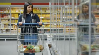 Genç bir kadın elinde alışveriş arabasıyla süpermarkete doğru yürür, raftaki eşyaları seçer ve onları el arabasına koyar. Alışveriş merkezindeki bir müşterinin portresi. Yüksek kalite 4k görüntü