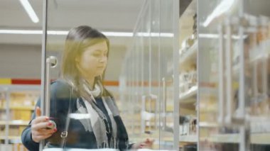 Kız yaklaşır ve süpermarketteki soğutmalı vitrinin kapılarını açar ve rafta duran malları seçer. Bir kadın süpermarkette alışveriş yapıyor. Yüksek kalite 4k görüntü