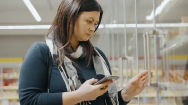 Akıllı telefonlu bir kız süpermarkette alışveriş arabasıyla gezer, cep telefonu ekranındaki malların listesine bakar ve alışveriş yapar. Ön planda. Yüksek kalite 4k görüntü