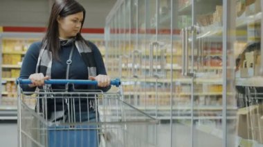 Elinde el arabası olan bir kız vitrine gider ve orada taze peynir seçer, kompozisyonu okuduktan sonra, bir seçim yapar ve malları el arabasına koyar. Süpermarkette alışveriş kavramı.