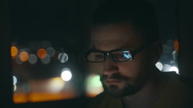 Gözlüklü genç bir adamın yüzü cep telefonunun ekranını aydınlatıyor. Adam sosyal ağda mesajlar yazıyor, Bokeh.4k 'teki şehir ışıklarının arka planındaki telefona odaklanıyor.