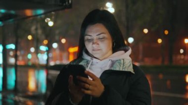 Geceleri neon ışıkları olan modern bir şehrin caddesinde duran güzel bir kadının portresi. Akıllı telefon kullanan ve şehir manzarasına bakan çekici bir kadın. Yüksek kalite 4k görüntü
