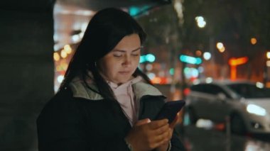 Geceleri neon ışıkları olan modern bir şehrin caddesinde duran güzel bir kadının portresi. Akıllı telefon kullanan ve şehir manzarasına bakan çekici bir kadın. Yüksek kalite 4k görüntü