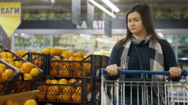 Kız süpermarketteki vitrindeki kutulara gider ve raftaki mandalinaları seçer. Bir kadın süpermarkette alışveriş yapıyor. 4k biçiminde yüksek kaliteli atış
