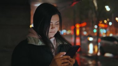 Geceleri neon ışıkları olan modern bir şehrin caddesinde duran güzel bir kadının portresi. Akıllı telefon kullanan ve şehir manzarasına bakan çekici bir kadın. Yüksek kalite 4k görüntü