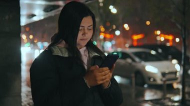 Geceleri neon ışıkları olan modern bir şehrin sokaklarında duran bir kızın portresi. Yağmur yağıyor. Islak şehir. Yağmurlu havalarda akıllı telefonlu bir kız. Yüksek kalite 4k görüntü