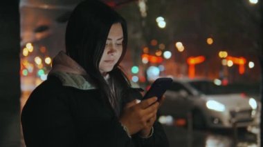 Geceleri neon ışıkları olan modern bir şehrin sokaklarında duran bir kızın portresi. Yağmur yağıyor. Islak şehir. Yağmurlu havalarda akıllı telefonlu bir kız. Yüksek kalite 4k görüntü