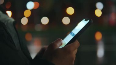 Bulanık gece ışıklarının arka planına karşı duran bir kız elinde bir telefon tutuyor ve parmaklarıyla haberlerde geziniyor. Cep telefonuyla haber okuyor. Yüksek kalite 4k görüntü