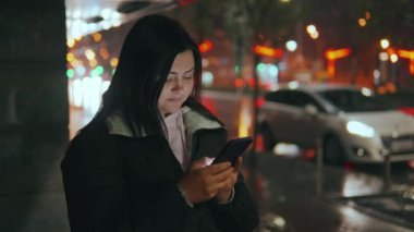 Geceleri neon ışıkları olan modern bir şehrin caddesinde duran güzel bir kadının portresi. Akıllı telefon kullanan ve şehir manzarasına bakan çekici bir kadın. Yüksek kalite 4k görüntü