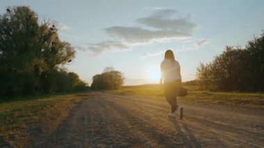 Tarlanın yakınındaki toprak yolda yürüyen bir kızın silueti. Kız yürüyor, ayaklarına bakıyor. Güzel bir akşam. Arka planda gün batımı. Yüksek kalite 4k görüntü