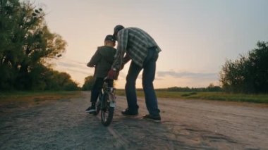 Mutlu aile, büyükbaba torununa gün batımında parkta bisiklet sürmeyi öğretiyor. Küçüklerin dedesi sokakta oynuyor. Yüksek kalite 4k görüntü