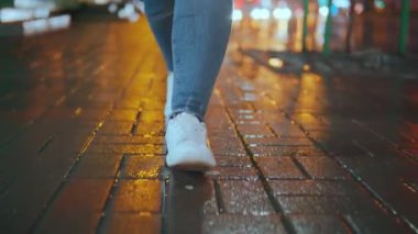 Yağmurdan sonra ıslak bir caddede, gece lambaları ve ıslak bir kaldırım taşının üzerinde parlayan farlarla tanınmayan bir yaya. Ayaklar kameraya doğru yürüyor. Yağmurdan sonra yürü. Yüksek kalite 4k görüntü