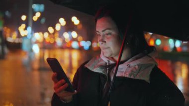 Modern bir şehrin caddesinde duran güzel bir kızın portresi. Geceleri sokak ışıklarıyla bir şemsiye altında. Çekici bir kadın akıllı telefon 45 'liği kullanarak haberlere bakıyor.