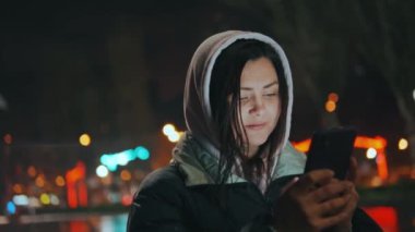 Geceleri neon ışıkları olan modern bir şehrin sokaklarında duran bir kızın portresi. Yağmur yağıyor. Islak şehir. Yağmurlu havalarda akıllı telefonlu bir kız. Yüksek kalite 4k görüntü