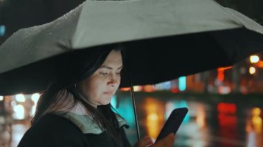 Modern bir şehrin caddesinde duran güzel bir kızın portresi. Geceleri sokak ışıklarıyla bir şemsiye altında. Çekici bir kadın akıllı telefon 45 'liği kullanarak haberlere bakıyor.