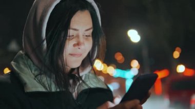 Geceleri neon ışıkları olan modern bir şehrin sokaklarında duran bir kızın portresi. Yağmur yağıyor. Islak şehir. Yağmurlu havalarda akıllı telefonlu bir kız. Yüksek kalite 4k görüntü
