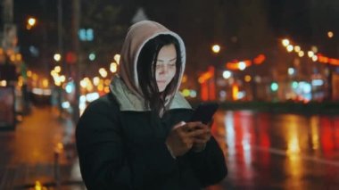 Modern bir şehrin caddesinde gece renkli ışıkları olan bir kadın. Akıllı telefon kullanan bir kadın şehir manzarasının arka planına bir mesaj yazıyor. 4K formatında çekim yapıyor.