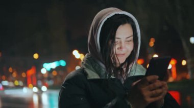 Kapüşonlu bir kız sosyal ağlarda cep telefonuyla iletişim kuruyor, yağmurdan sonra gece sokakta duruyor. Cep telefonuyla sohbet etmek. Yüksek kalite 4k görüntü
