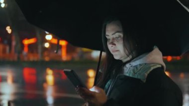 Modern bir şehrin caddesinde duran güzel bir kızın portresi. Geceleri sokak ışıklarıyla bir şemsiye altında. Çekici bir kadın akıllı telefon 45 'liği kullanarak haberlere bakıyor.