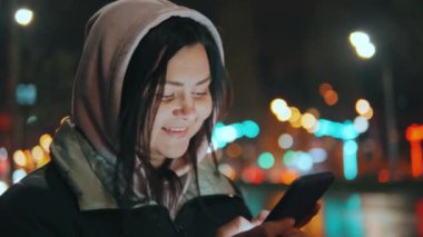 Kapüşonlu bir kız sosyal ağlarda cep telefonuyla iletişim kuruyor, yağmurdan sonra gece sokakta duruyor. Cep telefonuyla sohbet etmek. Yüksek kalite 4k görüntü