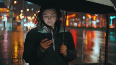 Modern bir şehrin caddesinde duran güzel bir kızın portresi. Geceleri sokak ışıklarıyla bir şemsiye altında. Çekici bir kadın akıllı telefon 45 'liği kullanarak haberlere bakıyor.