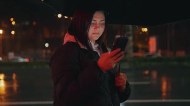 Bir kadın gece bulanık bir yol lambası deliğinde cep telefonuyla iletişim kuruyor. Yağmurdan sonra şehirde akıllı telefon kullanarak iletişim. Yüksek kalite 4K görüntü.