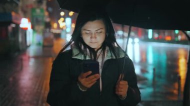 Genç bir kadın yağmurlu bir gecede dışarıda yürürken elinde şemsiye tutarken akıllı telefon kullanıyor ve ekranla yüzünü aydınlatıyor. Yağmurlu bir akşam.Yüksek kalite 4K görüntü.