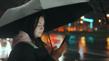 Modern bir şehrin caddesinde duran güzel bir kızın portresi. Geceleri sokak ışıklarıyla bir şemsiye altında. Çekici bir kadın akıllı telefon 45 'liği kullanarak haberlere bakıyor.