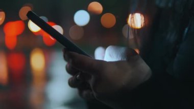 Bir kadın gece bulanık bir yol lambası deliğinde cep telefonuyla iletişim kuruyor. Yağmurdan sonra gece şehrinde akıllı telefon kullanarak iletişim. 4k biçiminde yüksek kaliteli atış