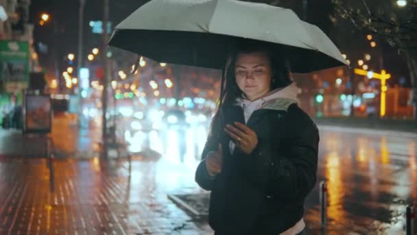 道路灯のぼんやりとしたボケの中で 夜に携帯電話を使って通信する女性 雨の後の夜の街でスマートフォンを使ったコミュニケーション 高品質4K映像 — ストック動画