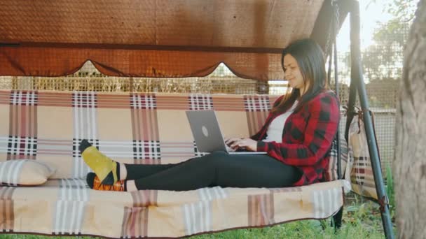 年轻的成年妇女在自家花园里用笔记本电脑干活 自由职业者坐在自家后院舒适的秋千上 一边用笔记本电脑 高质量的4K镜头 — 图库视频影像