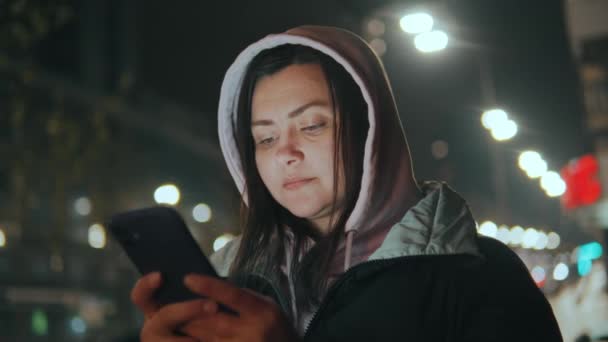 雨の降る夜の街の少女を背景に 夜に路上に立つ少女は メッセージと笑顔を書き込みます 携帯電話を使ったソーシャルネットワークでのコミュニケーション高品質4K映像 — ストック動画