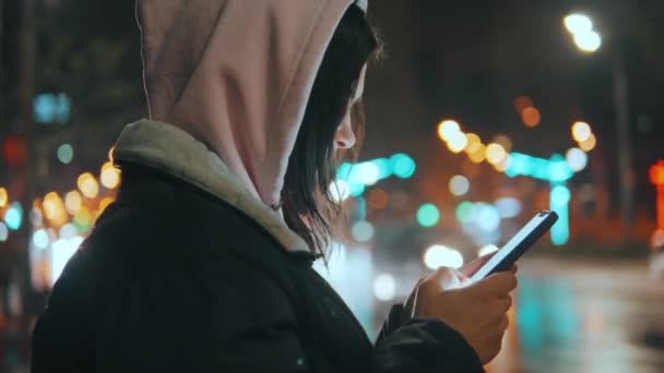 一个美丽的女人站在现代城市街道上的画像 夜晚有霓虹灯 迷人的女人用智能手机环视城市风景 高质量的4K镜头 — 图库视频影像