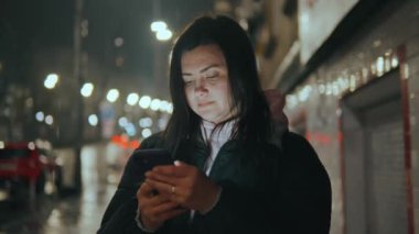 Gece vakti yağmurlu bir şehir kızının arka planına karşı duran kız bir mesaj yazar ve gülümser. Cep telefonu kullanılarak sosyal ağlarda iletişim. Yüksek kalitede 4K görüntü.