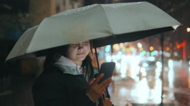 Geceleri neon ışıkları olan modern bir şehrin sokaklarında duran bir kızın portresi. Yağmur yağıyor. Islak şehir. Yağmurlu havalarda akıllı telefonlu bir kız. Yüksek kalite 4k görüntü