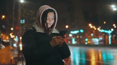 Kapüşonlu bir kız sosyal ağlarda cep telefonuyla iletişim kuruyor, yağmurdan sonra gece sokakta duruyor. Cep telefonuyla sohbet etmek. Yüksek kalite 4k görüntü