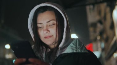 Genç bir kız, gece vakti şehrin arka planında duran kapüşonlu arabaların ışıklarına karşı, yağmurdan saklanıyor ve cep telefonuna bir mesaj yazıyor.