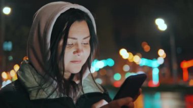 Geceleri neon ışıkları olan modern bir şehrin sokaklarında duran bir kızın portresi. Yağmur yağıyor. Islak şehir. Yağmurlu havalarda akıllı telefonlu bir kız. Yüksek kalite 4k görüntü