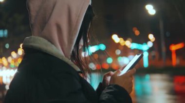 Sokakta duran modern şehir kızı sokak lambalarıyla dolu güzel bir bokeh 'le gece manzarasının arka planında akıllı telefonlu bir kızın yan görüntüsü. 4k