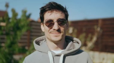 Çekici bir gencin portresi. Güneş gözlüklü bir adam kameraya mutlu bir şekilde bakar ve güneş ışınlarının tadını çıkarırken gülümser. Yüksek kalite 4k görüntü