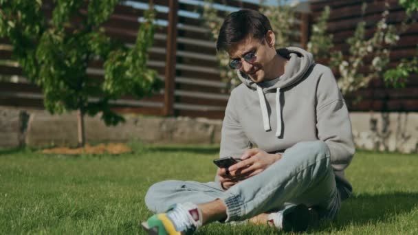 一个戴着太阳镜 手里拿着手机的帅哥坐在院子里的草坪上 微笑着看着摄像机 通过社交网络进行交流 约会网站 高质量的4K镜头 — 图库视频影像