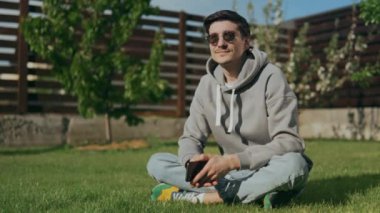 Güneş gözlüklü neşeli bir adam elinde telefonla bahçede çimlerin üzerinde oturuyor, gülümsüyor ve gökyüzüne bakıyor, hayatın tadını çıkarıyor. 4k biçiminde yüksek kaliteli atış