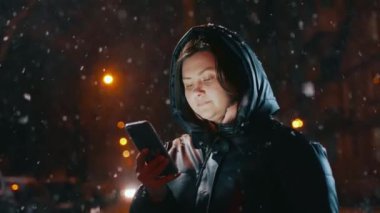 Elinde cep telefonu olan güzel bir kız fenerlerle aydınlatılmış bir kış caddesinde duruyor ve telefon ekranına bakıyor. Kar yağıyor. Sosyal ağlarda iletişim. Yüksek kalite 4k görüntü