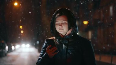 Elinde cep telefonu olan güzel bir kız fenerlerle aydınlatılmış bir kış caddesinde duruyor ve telefon ekranına bakıyor. Kar yağıyor. Sosyal ağlarda iletişim. Yüksek kalite 4k görüntü