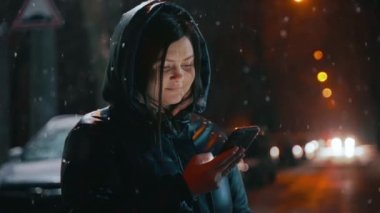 Elinde cep telefonu olan güzel bir kız fenerlerle aydınlatılmış bir kış caddesinde duruyor ve telefon ekranına bakıyor. Kar yağıyor. Sosyal ağlarda iletişim. Yüksek kalite 4k görüntü