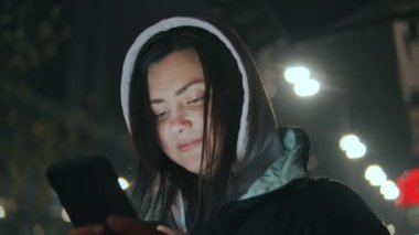 Geceleri neon ışıkları olan modern bir şehrin caddesinde duran güzel bir kadının portresi. Akıllı telefon kullanan ve şehir manzarasına bakan çekici bir kadın. Yüksek kalite 4k görüntü