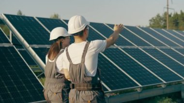Güneş enerjisi santralinde yürüyen üniformalı kadın ve erkek mühendislerin yan görüntüsü. İşçiler istasyonun işleyişini tartışıyor. Günümüz güneş panelleri. Güneş istasyonlarının gelişimi için kavram.
